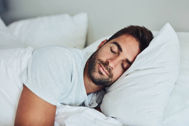Hombre con barba durmiendo en una cama con sabanas y colcha blanca apunto de despertarse